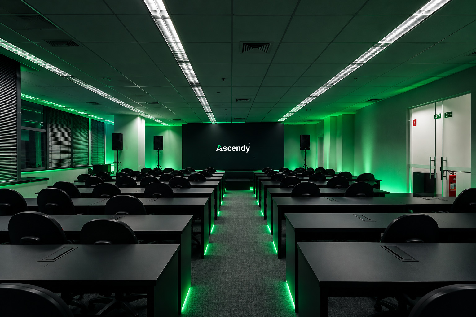 Sala da imersão montada em formato de sala de aula, com mesas pretas, iluminação verde e o telão com a marca Ascendy